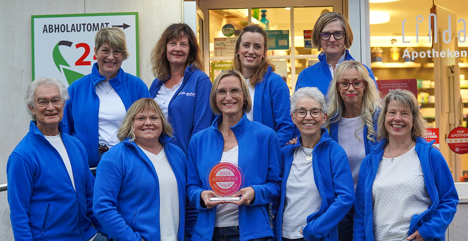 Eine Gruppe von zehn Personen stehen vor der Eifel Apotheke und lächelt in die Kamera. Alle tragen blaue Jacken über weißen Oberteilen. Eine Person in der Mitte hält eine rote Auszeichnung mit der Aufschrift „2025 Apotheke – der neuen Zeit“. Im Hintergrund ist der Eingang der Apotheke zu sehen, mit einem Schild „LINDA Apotheken“ und einem Hinweis auf einen 24-Stunden-Abholautomaten.